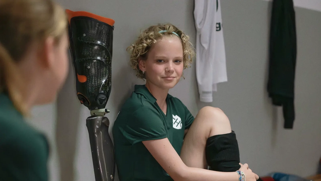 In einer Umkleidekabine sitzen zwei Mädchen in grünen Trikots. Eine von beiden schaut in die Kamera und hat ihr linkes Bein angewinkelt vor sich auf der Bank abgestellt. Am rechten Bein trägt sie eine Sportprothese.