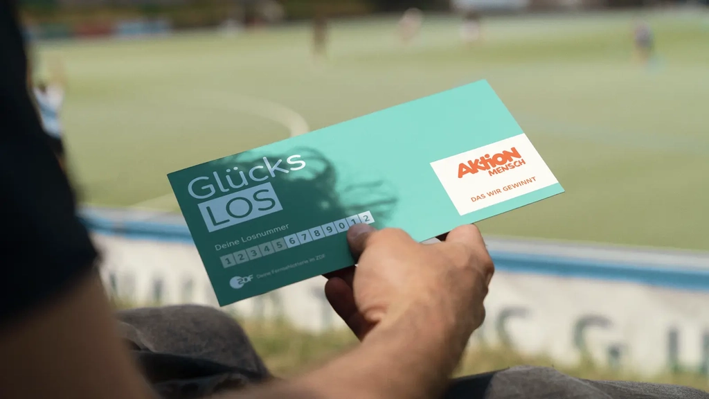Eine Hand hält ein Glücks-Los der Aktion Mensch in die Luft. Im Hintergrund sieht man einen Hockeyplatz.