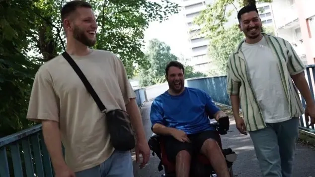 Drei junge Männer sind draußen unterwegs. Einer von ihnen benutzt einen Elektrorollstuhl. Sie unterhalten sich miteinander und lachen.