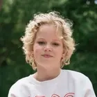 Ein blondes Mädchen im weißen Sweatshirt sitzt auf einer Holzbank. Im Hintergrund sind Bäume zu sehen. Sie hat ihre Unterschenkel überkreuzt. Rechts trägt sie eine Beinprothese.