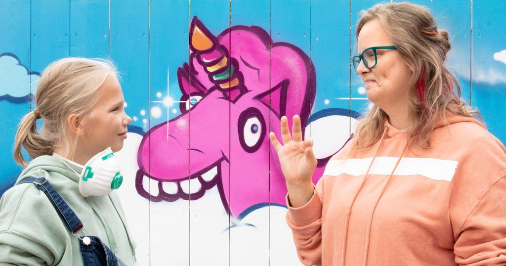 Die gehörlose Sylvia steht mit ihrer Tochter Lea vor einer Wand, auf der ein buntes Graffiti von einem Einhorn zu sehen ist.