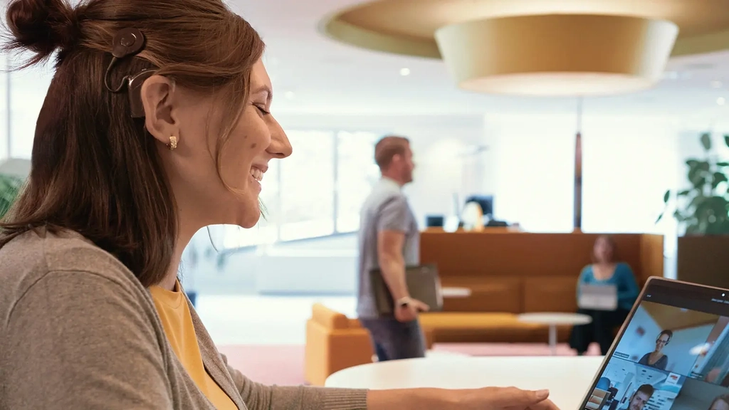 Eine junge Frau mit einem Cochlea-Implantat sitzt vor einem Laptop und nimmt an einer Videokonferenz teil.