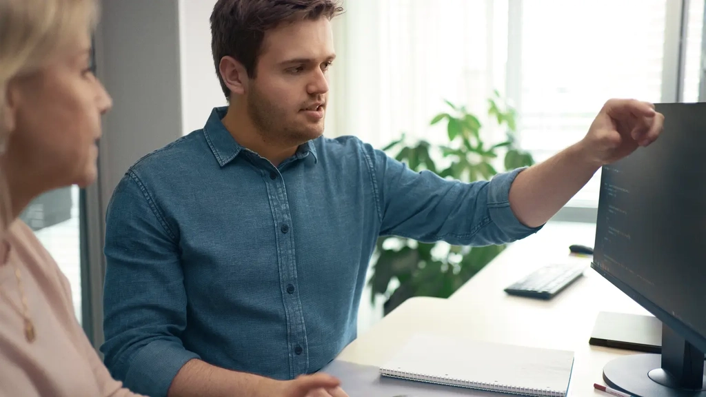Ein junger Mann und eine Frau sitzen nebeneinander vor einem Bildschirm. Der junge Mann zeigt mit der linken Hand auf den Monitor.