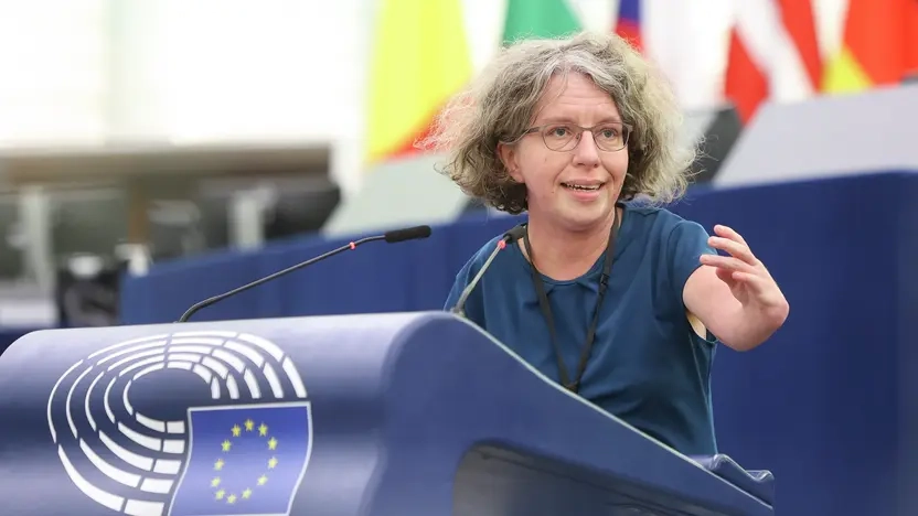Eine Frau mit gewellten ergrauenden Haaren, Brille, dunkelblauer Bluse und kurzen Armen steht am Rednerpult des EU-Parlaments