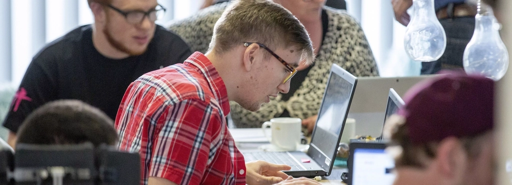Ein junger Mann arbeitet an einem Laptop. Um ihn herum sitzen weitere Personen mit und ohne Behinderung und arbeiten ebenfalls.