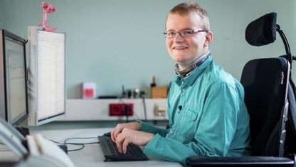 Ein junger Mann sitzt im Rollstuhl am Schreibtisch. Seine Hände liegen auf der Tastatur, er lächelt seitlich in die Kamera.