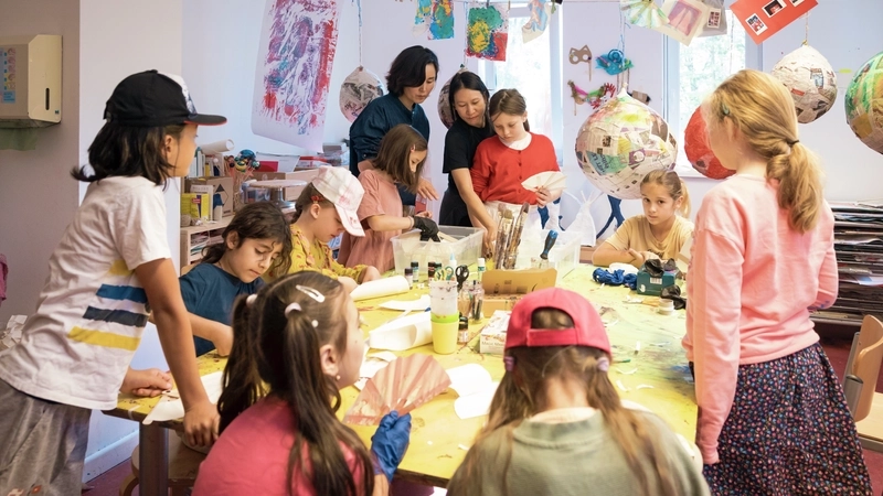 Eine multikulturelle Gruppe aus etwa zehn Kindern im Grundschulalter, an einem großen Tisch basteln. Zwei erwachsene Frauen geben einem der Mädchen gerade Tipps.  Auf dem Tisch stehen viele Bastelsachen. An einer Leine, die quer durch das Kunstatelier gespannt ist, hängen fertige Kunstwerke.