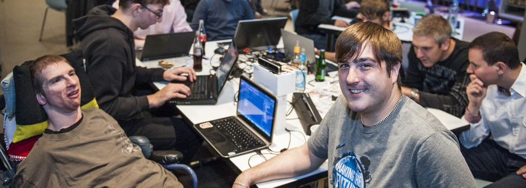 Ein Mann im Rollstuhl und ein Mann ohne Behinderung lächeln in die Kamera. Im Hintergrund arbeiten Menschen an Laptops.