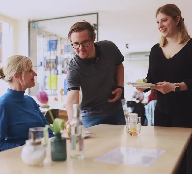 Zwei Frauen sitzen an einem Holztisch. Ein Mann mit Down-Syndrom und eine junge Frau servieren jeder einen Teller mit Kuchen.