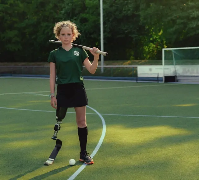 Annika, ein Mädchen mit lockigem Haar und Beinprothese steht auf einem Hockeyplatz. Zwischen ihren Beinen liegt der Ball, ihren Schläger hat sie lässig über die Schultern gelegt.