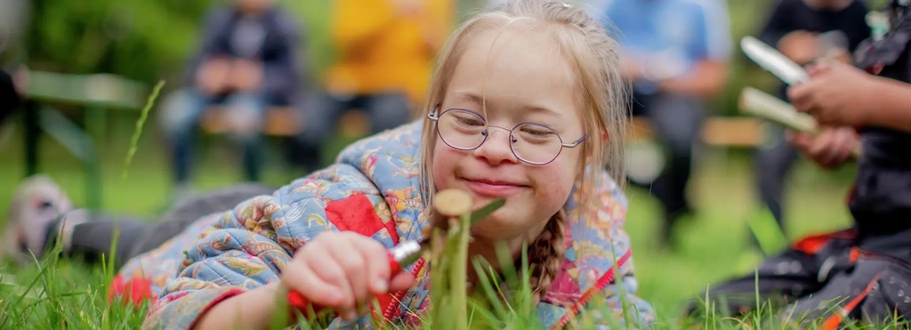 Auf einer Wiese liegt ein Mädchen mit Brille und schneidet eine Blume ab. Im Hintergrund kann man unscharf weitere Kinder sehen.