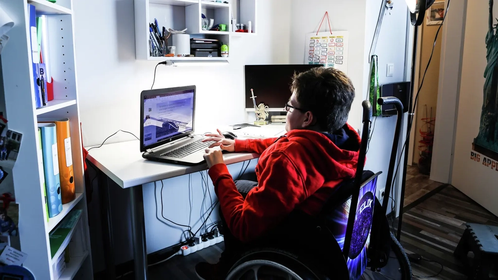 Ein junger Mann im Rollstuhl sitzt an einem Schreibtisch und bedient einen Laptop.