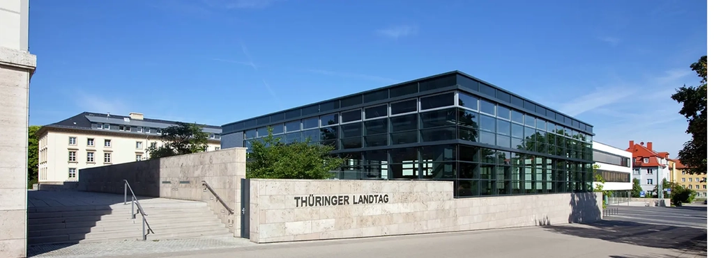 Landtagsgebäude in moderner Glasarchitektur auf hellem Steinpodest, an dessen Wand der Schriftzug Thüringer Landtag zu sehen ist