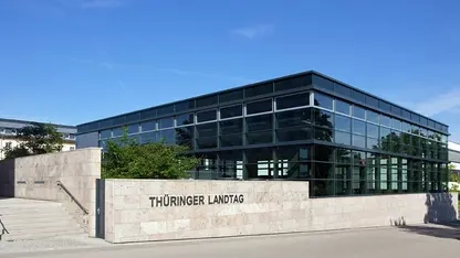 Landtagsgebäude in moderner Glasarchitektur auf hellem Steinpodest, an dessen Wand der Schriftzug Thüringer Landtag zu sehen ist