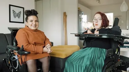 Zwei junge Frauen im E-Rolli sitzen nebeneinander in einer Wohnung