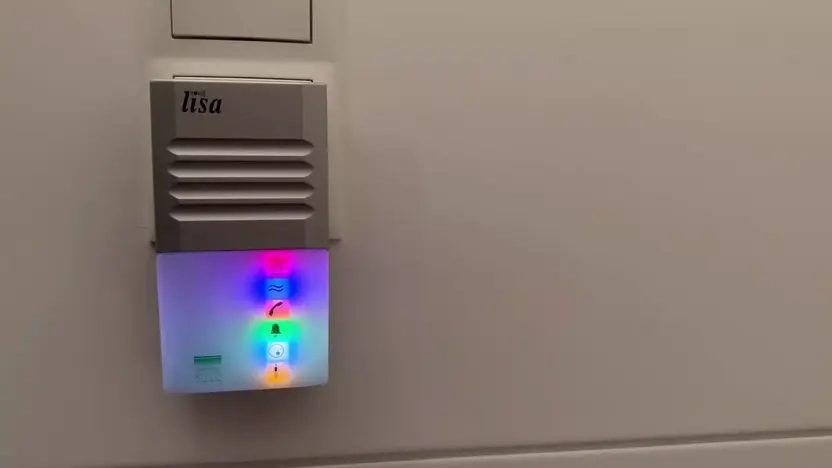 Ein flaches rechteckiges Gerät steckt in einer Steckdose. Im unteren Teil sind verschiedene Symbole zu sehen für Tür, Rauchmelder, Telefon und schreiendes Kind. Für jedes der Symbole gibt es ein unterschiedliches Lichtsignal.