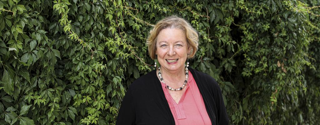 Eine Frau mit mittellangem, blonden Haar steht lachelnd vor einer Hecke.