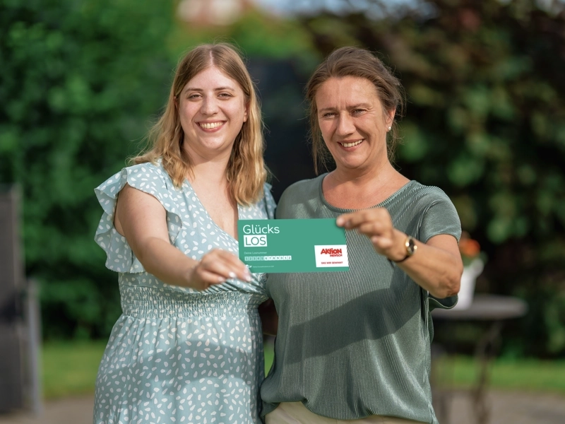 Zwei Frauen stehen lächelnd nebeneinander im Garten und halten gemeinsam ein Glücks-Los hoch.