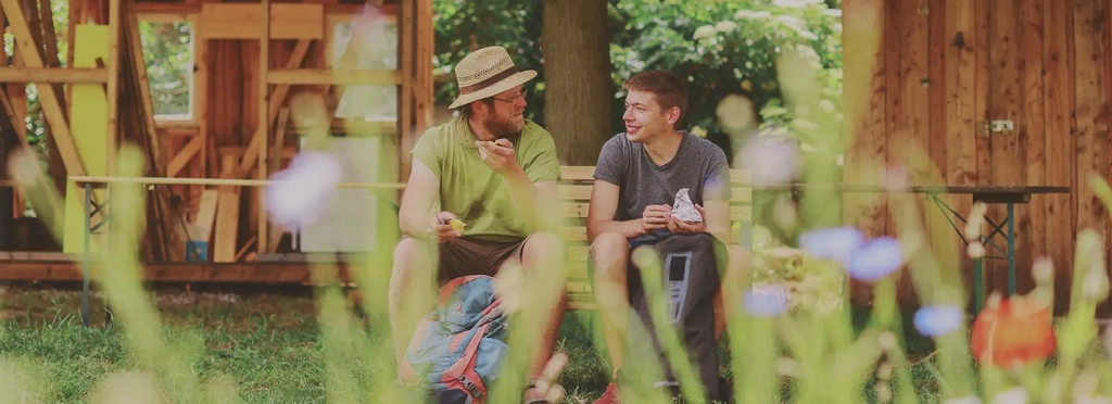 Ein Mann mit Hut und ein Jugendlicher sitzen auf einer Bank im Grünen und essen ein Butterbrot.