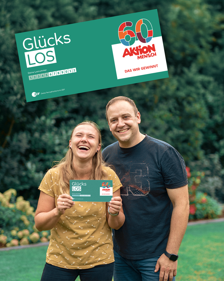 Die Gewinner*innen Bozena und Sebastian stehen in sommerlicher Kleidung nebeneinander in ihrem Garten. Bozena hält ihr Glückslos in die Kamera und lacht. Über den beiden ist groß das Jubiläumslos der Aktion Mensch abgebildet. Es ist ein Glücks-Los, auf dem eine große bunte 60 zu sehen ist.