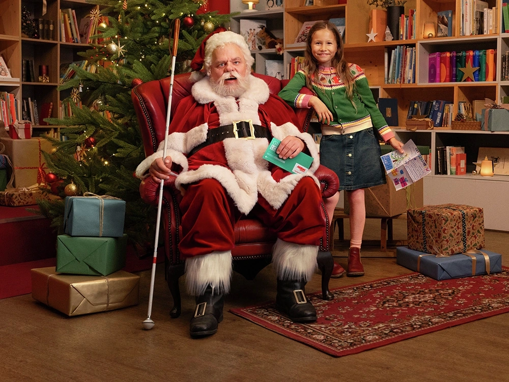 Ein Mann in rot-weißem Weihnachtsmannkostüm sitzt in einem roten Ohrensessel. Im Hintergrund steht ein geschmückter Tannenbaum, drumherum sind Geschenke verteilt. Der Weihnachtsmann hält in seiner rechten Hand einen Blindenstock, in der linken ein Glücks-Los der Aktion Mensch. Neben ihm steht ein junges Mädchen und hält einen Wunschzettel in der Hand. Beide schauen freundlich zur Kamera und lächeln.