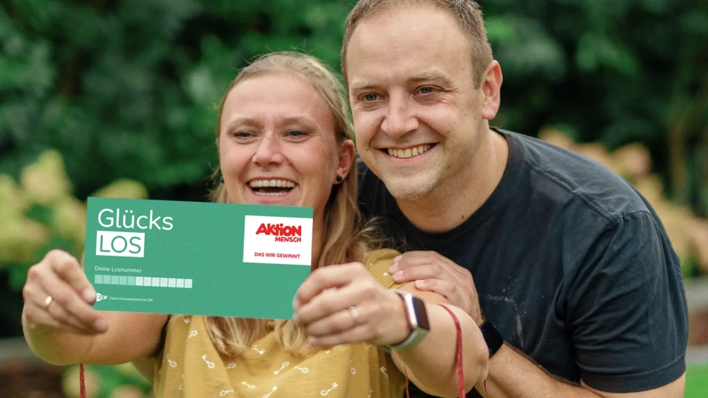 Die Gewinner*innen Bozena und Sebastian stehen in sommerlicher Kleidung nebeneinander in ihrem Garten. Bozana hält ihr Glückslos in die Kamera. Beide lachen.