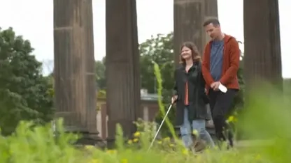 Eine junge Frau mit Blindenstock und ein junger Mann gehen spazieren. Im Vordergrund ist Wiese zu sehen. Im Hintergrund Säulen der Universität Potsdam.