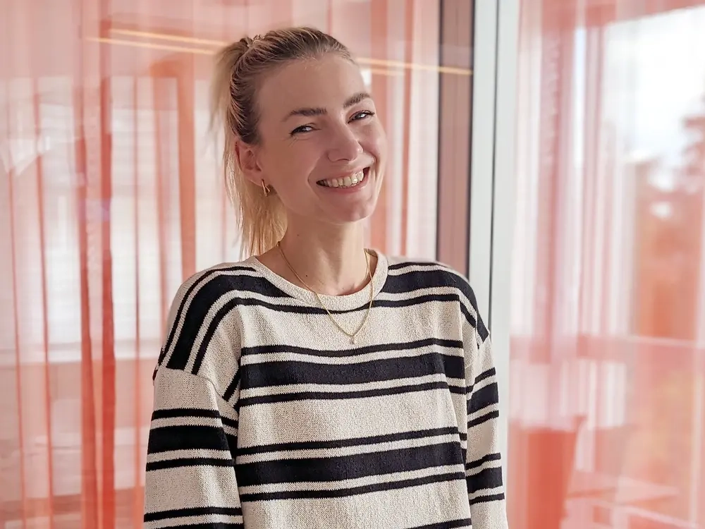 Halbtotale einer jungen Frau in einem blau weiß gestreiften Pulli, die vor einer Glaswand mit Vorhängen steht. Sie lacht in die Kamera. Ihre langen blonden Haare sind zu einem Pferdeschwanz gebunden.