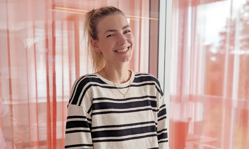 Halbtotale einer jungen Frau in einem blau weiß gestreiften Pulli, die vor einer Glaswand mit Vorhängen steht. Sie lacht in die Kamera. Ihre langen blonden Haare sind zu einem Pferdeschwanz gebunden.