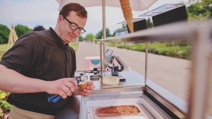 Ein junger Mann mit Brille steht hinter einem Eismobil und gibt eine Kugel Eis auf eine Waffel.
