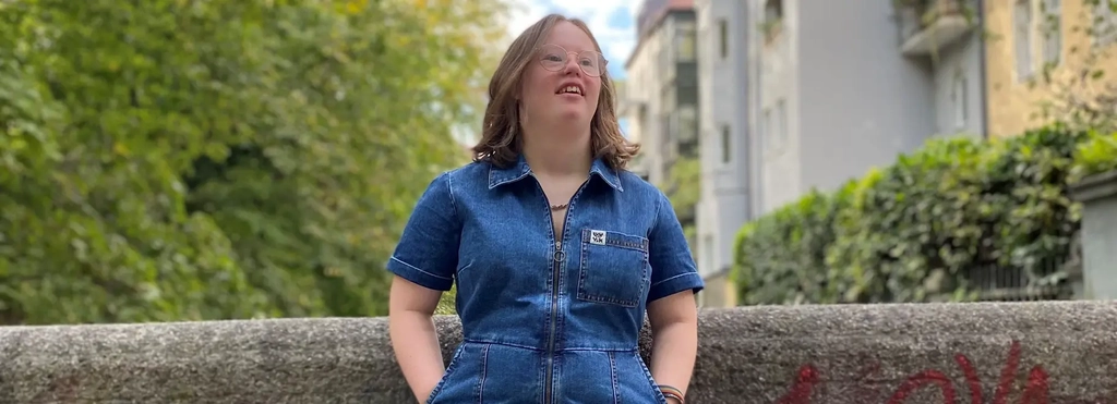 Eine junge Frau mit Down-Syndrom lehnt lässig an einer Mauer. Sie hat schulterlange blonde Haare, trägt eine Brille und einen blauen Hosenanzug aus Jeansstoff, und Sneaker mit Leopardenfell-Muster. Im Hintergrund Hausfassaden und Bäume.