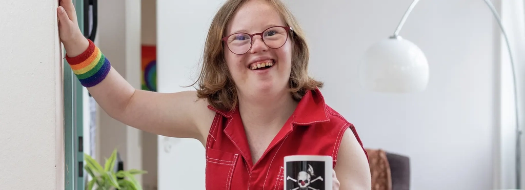 Natalie Dedreux steht in einem roten Jumpsuite in einem Wohnzimmer und lacht in die Kamera. Sie hält sich mit der rechten Hand an der Wand fest.