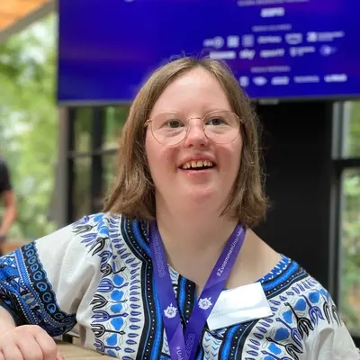 Eine junge Frau mit Down-Syndrom an einem Stehtisch. Vor ihr liegen Moderationskarten und ein Mikrophon. Sie trägt ein blau-weiß gemustertes afrikanisch anmutendes Oberteil an dem ein Schild mit ihrem Namen angebracht ist.