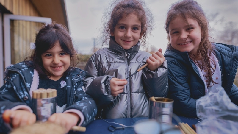 Drei kleine Mädchen in dicken Jacken posieren stolz an einem Tisch im Freien, an dem sie gerade Bienenhotels bauen.