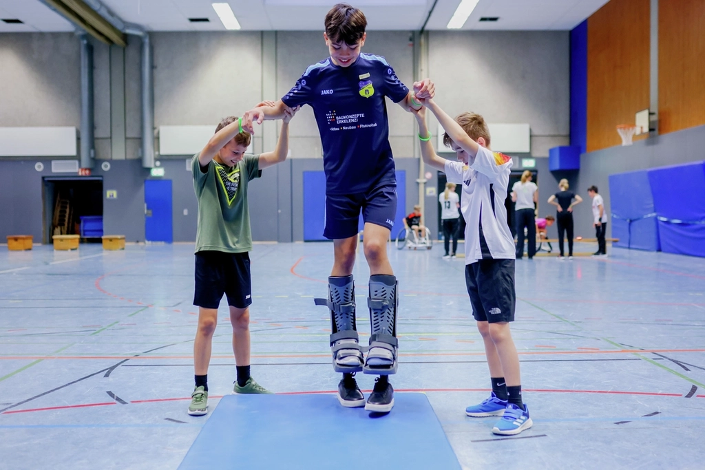 Ein Junge probiert aus, wie es sich auf Prothesen gehen lässt. Zwei Mitschüler stützen ihn links und rechts, damit er das Gleichgewicht halten kann.