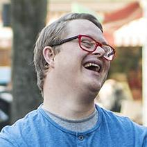 Junger Mann mit Down-Syndrom. Er trägt eine rote Brille und lacht.