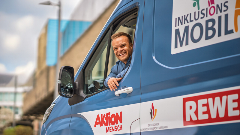 In einem blauen Kleintransporter sitzt ein kleinwüchsiger, sportlicher Mann mit kurzen blonden Haaren. Er sieht aus dem Fenster der Fahrertür und lacht in die Kamera. Auf dem Bus sind Ausfkleber angebracht. Auf einem steht "Inklusionsmobil". Auf den anderen sind die Logos der kooperatiosnpartner zu sehen, die diesen Bus unterstützen: Aktion Mensch, Deutscher Behindertensportverband und REWE.