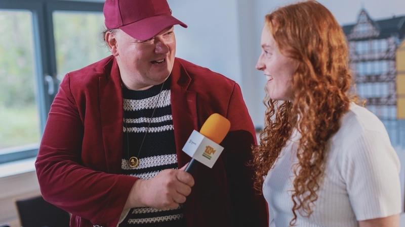 Ein großer Mann mit Sakko, grob gestrickten schwarz-weiß-gestreiftem Pulli und einem Cappy hält einer jungen Frau mit langen, gelockten roten Haaren und hellem geripptem T-Shirt ein Mikrofon hin, in das sie hineinspricht. Die beiden stehen in einem etwas kahlen Raum mit weißen Wänden.
