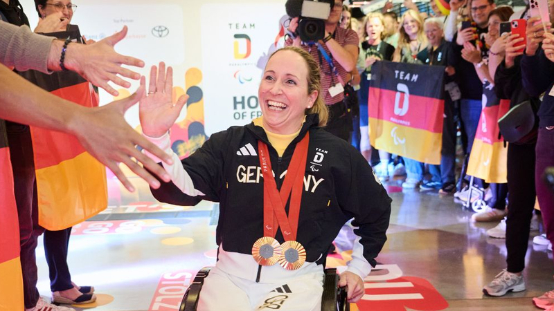 Para Radsportlerin Annika Zeyen-Giles fährt in ihrem Rollstuhl an einer jubelnden Menschmenge im Deutschen Haus Paralympics Paris 2024 entlang. Es werden Deutschlandfahnen geschwenkt. Die Rollstuhlfahrerin klatscht sich mit einem Mann ab