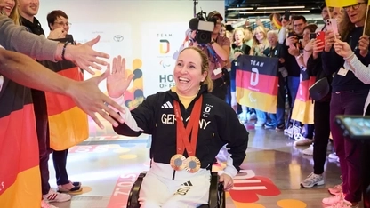Para Radsportlerin Annika Zeyen-Giles fährt in ihrem Rollstuhl an einer jubelnden Menschmenge im Deutschen Haus Paralympics Paris 2024 entlang. Es werden Deutschlandfahnen geschwenkt. Die Rollstuhlfahrerin klatscht sich mit einem Mann ab
