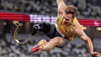 Eine Aufnahme vom Paralympionik Markus Rehm beim Hochsprung bei den Paralympics 2020 in Tokyo. Er ist mitten im Sprung und hat die Arme weit ausgebreitet. Am rechten Bein trägt er eine Prothese.
