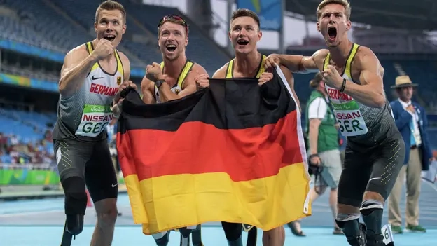 Die deutsche Mannschaft mit Markus Rehm, David Behre, Felix Streng und Johannes Floors gewinnt die Goldmedaille über 4x100m der Männer. Sie stehen dicht beieinander, halten die Deutschlandflagge und strahlen vor Freude.