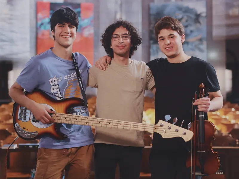 Drei Jugendliche stehen haben jeweils einen Arm und die Schulter des anderen gelegt. Sie halten eine E-Gitarre und eine Geige in der Hand.