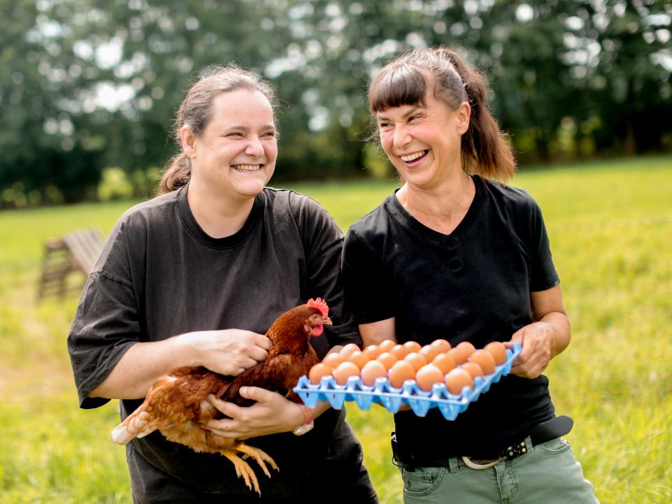 Zwei Frauen stehen nebeneinander und lachen. Die linke Frau hält ein Huhn auf den Arm. Und die Frau auf der rechte Seite eine Palette mit Eiern.