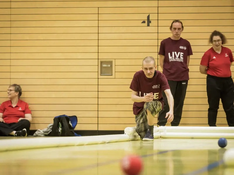 Ein Mann wirft in gebückter Haltung eine Bocciakugel auf das Spielfeld. Hinter ihm stehen zwei weitere Mitspielende.