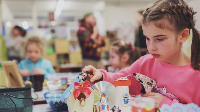 Ein braunhaariges Mädchen klebt mit einer Heißklebepistole Figuren auf ein kleines Haus aus Karton.