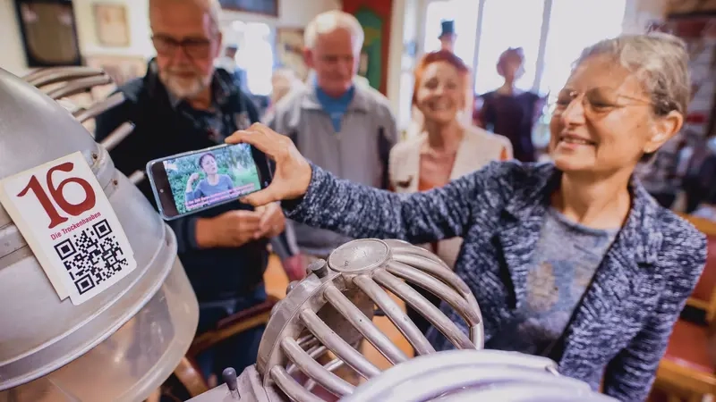 Eine Gruppe Besucher*innen steht an einem alten Frisierstuhl. Über ein Mobiltelefon schauen sie sich ein Gebärdensprachvideo zum Exponat an.