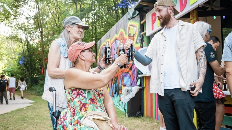 Eine Frau in Hippie-Kleidung und eine zweite Frau in Sommerkleid, die im Rolli sitzt, stoßen mit Colaflaschen mit einem tätowierter Mann mit Vollbart an. Alle drei tragen Baseball-Cappies. Sie stehen auf einem Festivalgelände vo reiner aus Holzbrettern zusammengezimmerten bunten Bar. Die Zugangswege zur Bar bestehen aus Konststoffelementen, die auf die Wiese gelegt sind.
