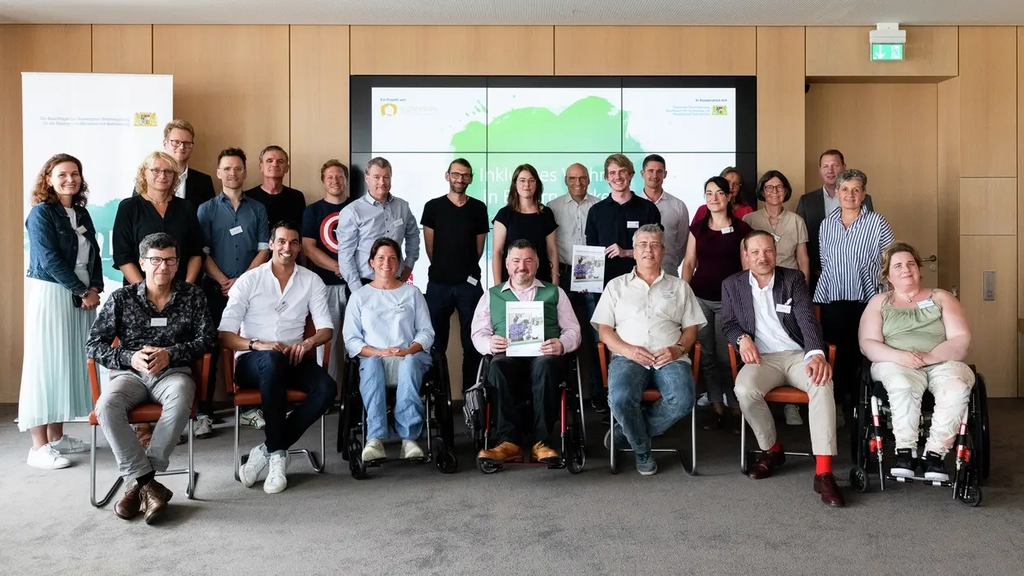 Eine Gruppe von etwa 25Frauen und Männern sitzen vor der Wand eines Konferenzraums und präsentieren eine Broschüre