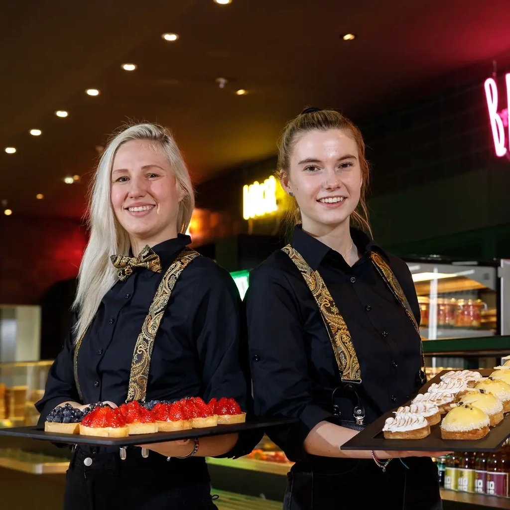 Zwei junge Frauen stehen nebeneinander in einem Bistro. Beide halten jeweils ein Tablett mit Törtchen in Händen.